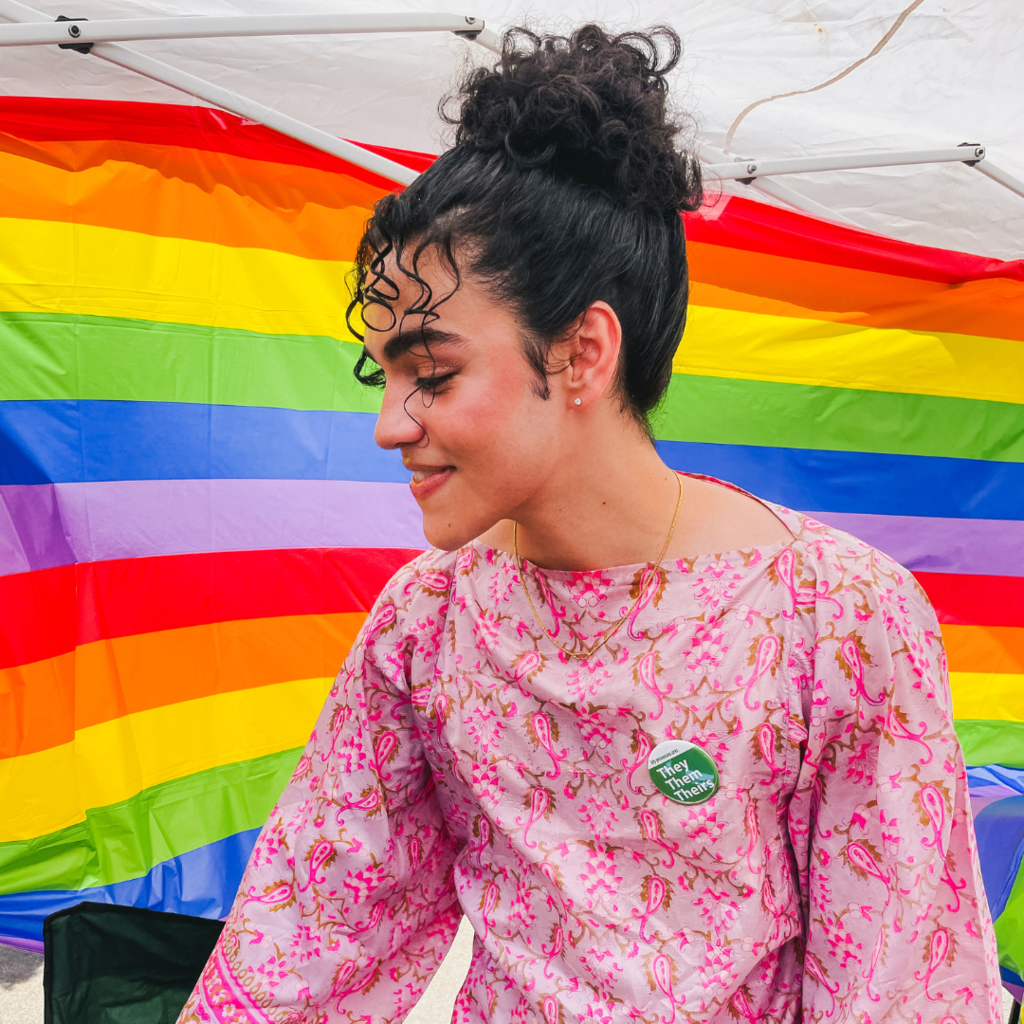 Paras is smiling off to the side while standing in front of a rainbow flag background.