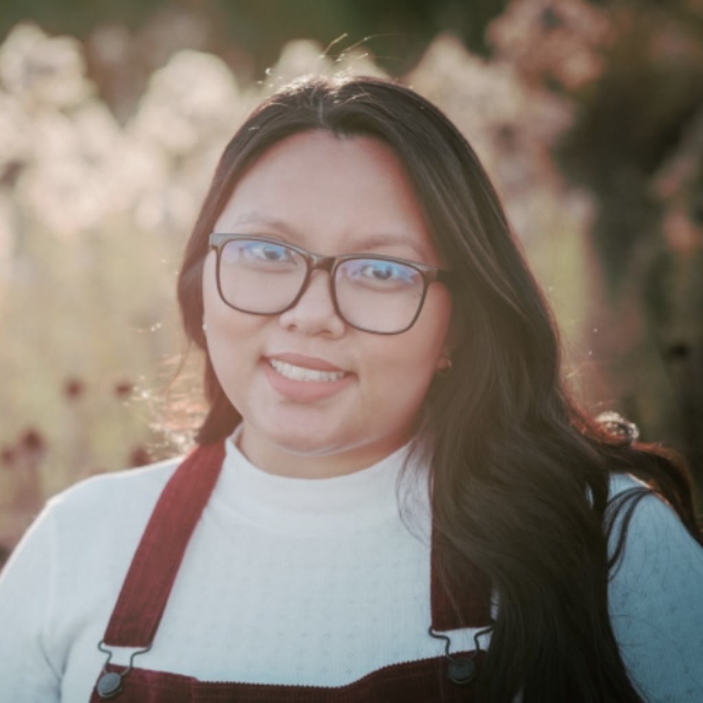 Linh is in front of a field of wheat. She is smiling into the camera.