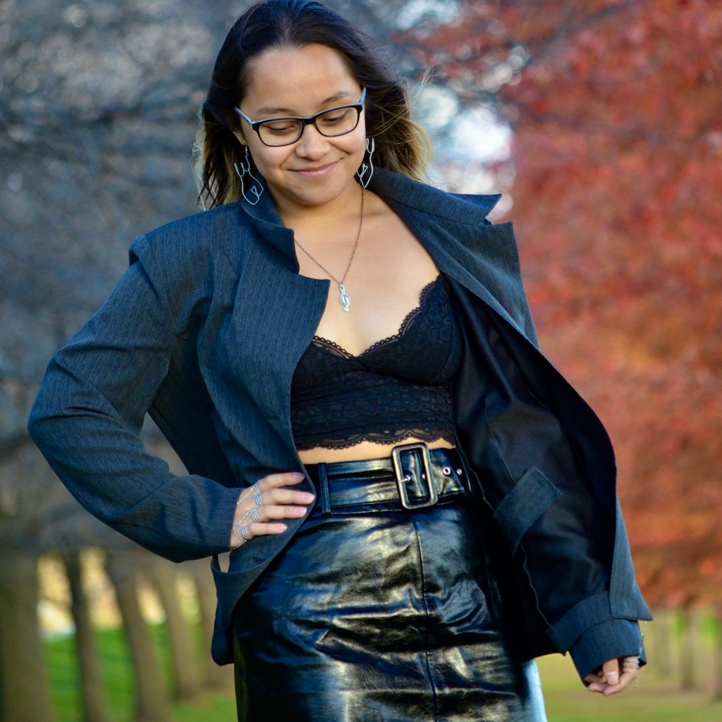 Fenix is glancing down and smiling, standing outside in front of autumn trees.