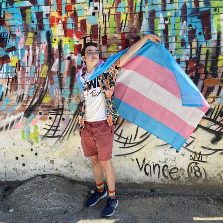 Logan is standing in front of a spray painted wall. They have a trans flag draped around their shoulders and are proudly holding it outward.