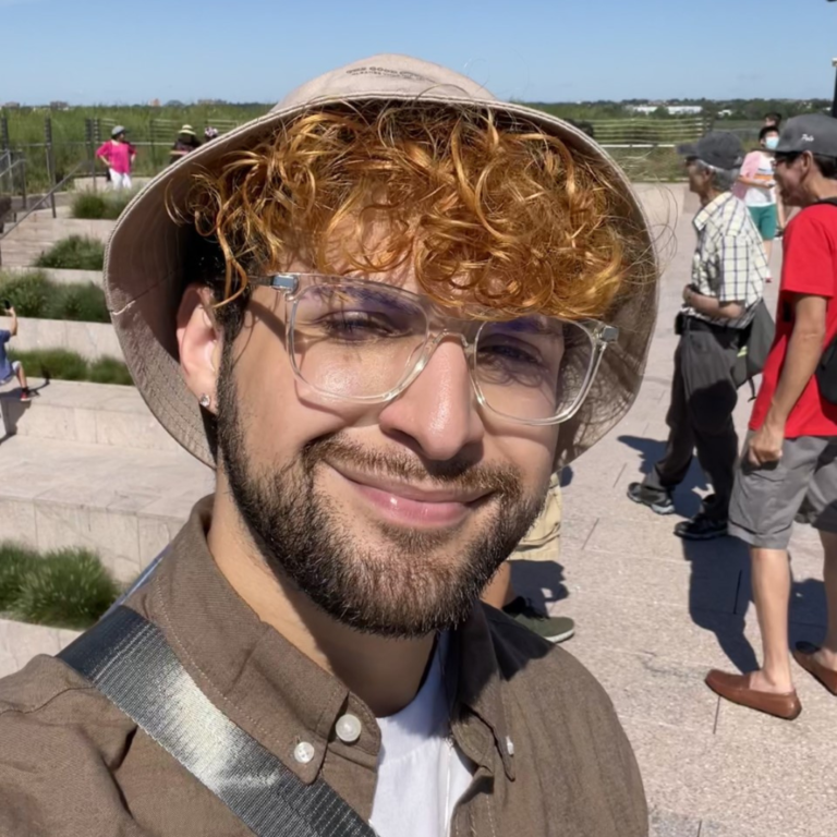 Joey is smiling into the camera in the sunlight. Joey is wearing transparent glasses and a bucket hat.